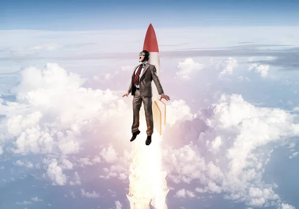 Businessman in suit and aviator hat flying on rocket in stratosphere. Superhero businessman flying with jetpack rocket in blue sky above clouds. Successful business startup. Career growth concept.