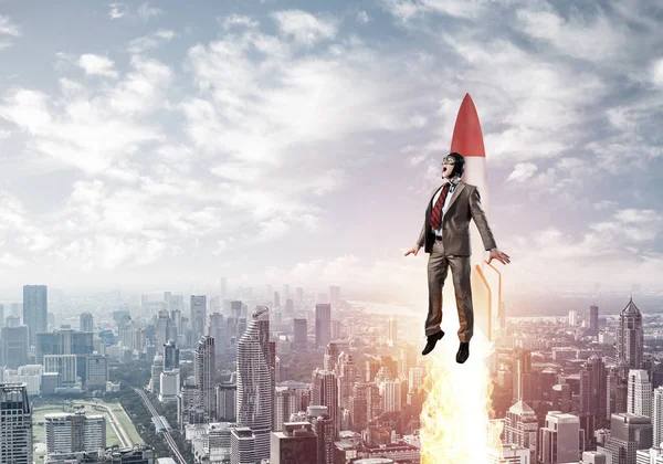 Businessman in suit and aviator hat flying on rocket. Superhero businessman flying with jetpack rocket in blue sky above modern downtown. Successful business startup. Career growth concept.