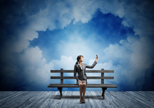 Young woman in business suit holding smartphone with raised hand. Girl with mobile phone sitting on wooden bench. Mobile marketing and digital technology. Beautiful skyscape with deep blue night sky