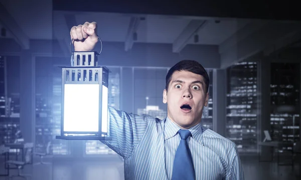 Horrified businessman holding glowing lantern on background office interior. Emotional corporate employee in shirt and tie looking for something at night. Shocked man with open mouth lost in dark