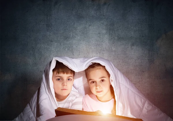 小女孩和男孩在毯子下看着对方 孩子们睡觉前在床上读魔法故事 年轻的妹妹和弟弟在睡衣一起在灰色的墙壁背景 — 图库照片