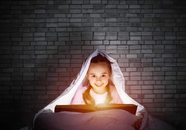 寝る前にベッドで本を読んで幸せな小さな女の子 毛布の下に隠れている懐中電灯を持つ美しい子供 壁を背景にパジャマ姿の笑顔の少女の肖像 おとぎ話を読む子供 — ストック写真