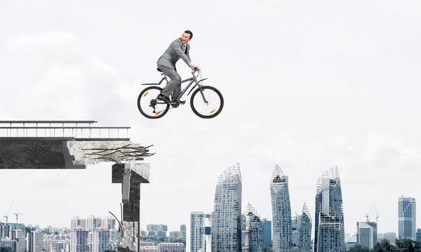 Businessman commuting to work by bike. Happy man wearing business suit riding bicycle in air. Handsome cyclist looking back on background modern downtown panorama and blue cloudy sky.