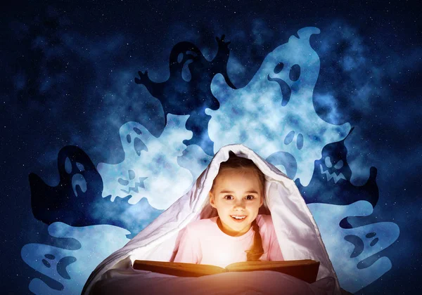 快乐的小女孩睡觉前在床上看书 手电筒藏在毯子下的孩子 穿着睡衣的微笑女孩和有趣的鬼魂回到夜空 儿童阅读魔法童话 — 图库照片