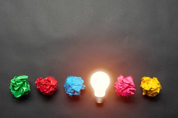 Lightbulb and crumpled colorful paper balls on black background. Successful solution of problem. Idea generation and brainstorming. Genius idea among failing ideas metaphor. Business motivation