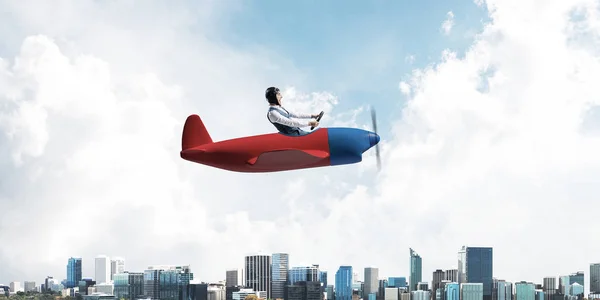 Businessman in aviator hat and goggles driving propeller plane above downtown with high modern buildings. Successful leader flying in small airplane. Blue sky landscape with clouds on background