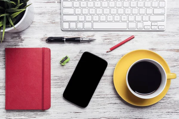 Top view businessman table with office accessories. Morning coffee time and planning new day. Flat lay wooden desk with computer keyboard, smartphone, cup of coffee, red diary and green plant in pot.