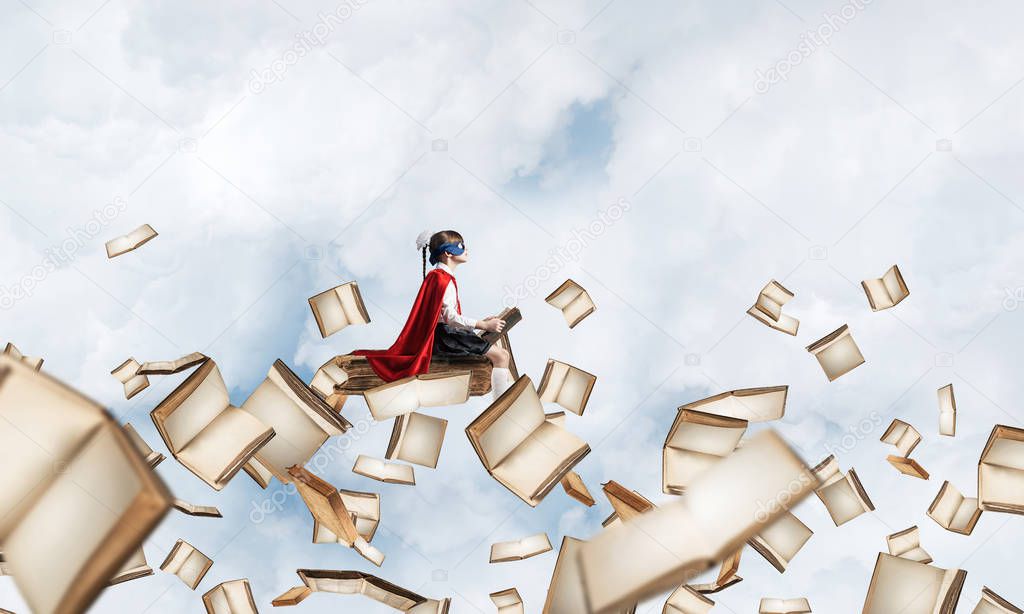 Little child in mask and cape floating on book in sky and reading