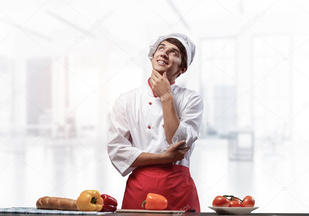 Young male chef standing with folded arms and looking up dreamily. Pensive culinary school student in white hat and red apron in light kitchen interior. Professional kitchen staff recruitment