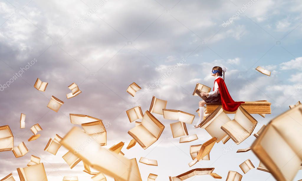 Little child in mask and cape floating on book in sky and reading