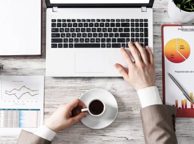 İnsan eliyle modern ofis çalışma alanı. Düz beyaz tahta masa, dizüstü bilgisayarlı klavye, iş bilgileri ve bir fincan kahve. Bilgisayarda çalışan işadamı elleri.