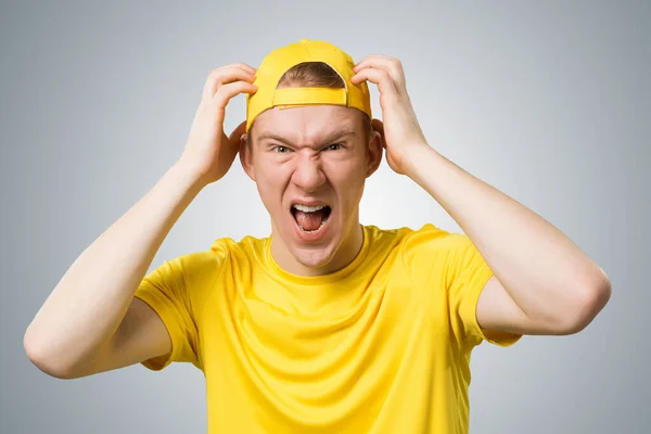 紧张的少年怒气冲冲地尖叫着 双手紧握着头 情绪激动的红头发男孩有恼怒的面部表情 穿着黄色T恤 头戴灰色背景棒球帽的人的画像 — 图库照片
