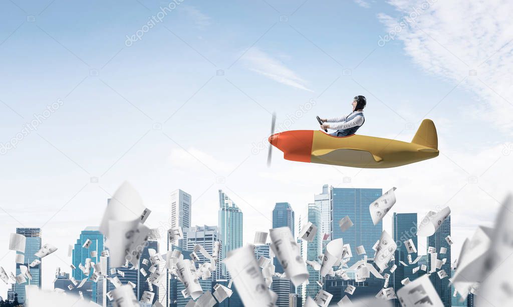 Businessman in aviator hat and goggles driving propeller plane above downtown. Papers falling down on background of skyscrapers. Funny man having fun in small airplane. Blue sky with clouds.