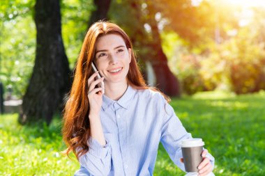 Genç kızıl saçlı kadın yaz parkında akıllı telefondan konuşuyor. Güneşli bir günde dışarıda dinlenen güzel bir kız. Yeşil çimlerin üzerinde oturan ve kahve içen sevimli, güler yüzlü kız. Kahve molası.