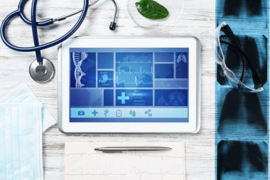Doktorların ofisinde laboratuvar hasta muayenesi. Ekranda tıbbi uygulama arayüzü olan tablet bilgisayar. Stetoskop, röntgen görüntüsü ve kardiyogram tahta masada. Tıbbi teşhis ve muayene