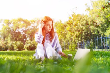 Genç kızıl saçlı kadın parkta yeşil çimlerde dizüstü bilgisayar kullanıyor. Gündelik giysiler içinde güzel bir kız güneşli bir yaz gününde dışarıda bilgisayarla çalışıyor. Serbest çalışma, blog yazma veya uzaktan çalışma kavramı.