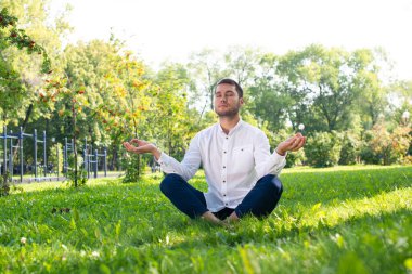 Genç adam nilüfer çiçeğiyle yeşil çimlerin üzerinde meditasyon yapıyor. Rahat giyinen yakışıklı adam yoga yaparken gözleri kapalıydı. Yaz günü dışarıda antrenman ve meditasyon. Sağlıklı yaşam tarzı ve rahatlama.