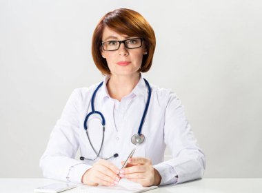 Ofiste oturan çekici kızıl saçlı doktor kameraya bakıyor. Beyaz önlüklü, steteskoplu beyaz arka planda duran terapist portresi. Profesyonel tıbbi teşhis ve tedavi.