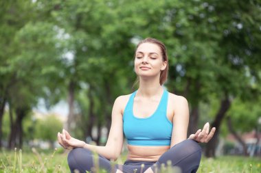 Kız, Lotus 'ta meditasyon yapıyor. Parktaki yeşil çimlerde poz veriyor. Gözleri kapalı barışçıl bir kadının portresi. Yaz günü dışarıda antrenman ve meditasyon. Doğayla uyum ve rahatlama. Yoga pratiği..