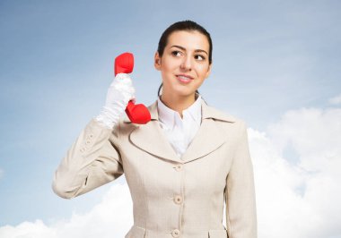 Kırmızı retro telefonu tutan çekici genç bir kadın. Beyaz takım elbiseli çağrı merkezi operatörü gökyüzü arka planında telefonla poz veriyor. Tele-pazarlama hattı. İş yardımı ve danışma.