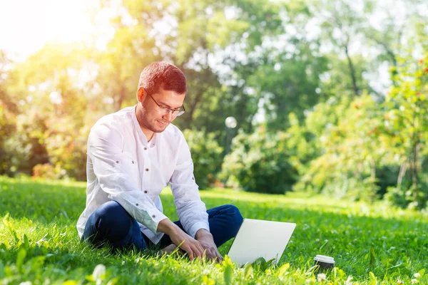 若いビジネスマンは緑の芝生の上に座って ラップトップコンピュータを使用しています 晴れた夏の日に公園でコンピュータを扱うハンサムな男 アウトドア自然の旅とリラクゼーション フリーランスの仕事のコンセプト — ストック写真