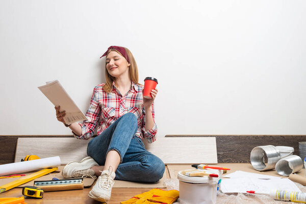 Interior designer studying renovation project. Smiling pretty girl sitting on floor with paper blueprint. Home remodeling and house interior redesign. Construction tools and materials for building.