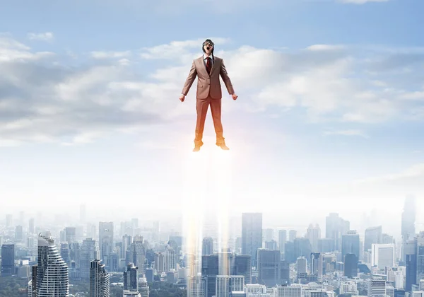 Businessman in suit and aviator hat flying in blue sky as superhero. Corporate manager as superman launching upwards with jet flame above modern city. Successful business startup motivation concept