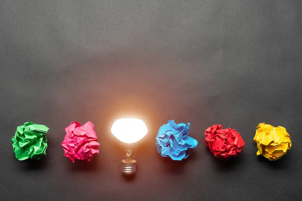 Lightbulb and crumpled colorful paper balls on black background. Successful solution of problem. Idea generation and brainstorming. Genius idea among failing ideas metaphor. Business motivation