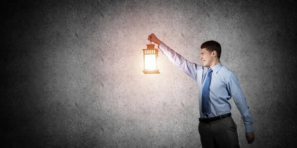 Happy businessman holding glowing lantern on background grunge wall. Side view of young man in shirt and tie looking for something in dark. Smiling business person lighting his way with lantern.