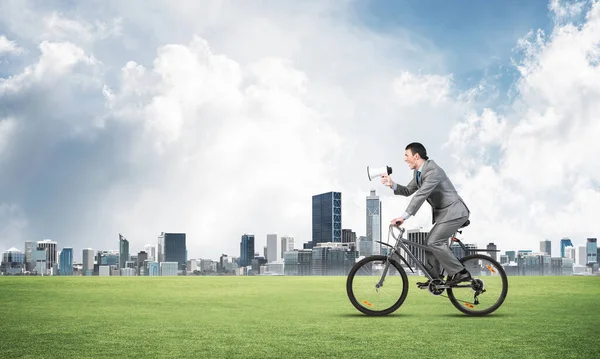 Businessman with megaphone on bike at sunny day. Marketing and advertising campaign. Manager in business suit riding bicycle on green meadow. Male cyclist on background of blue sky and city on horizon