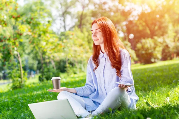 若い赤毛の女性は緑の芝生の上でノートパソコンとコーヒーカップで蓮のポーズで瞑想します 閉じた目でヨガの練習カジュアルウェアの美しい女の子 夏の日の屋外の仕事とリラクゼーション — ストック写真