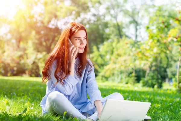 若い赤毛の女性がノートパソコンで緑の芝生の上に座っている 晴れた夏の日に公園でスマートフォンで話しているチャーミングな女の子 モバイルコミュニケーションと会話 屋外での仕事とリラクゼーション — ストック写真