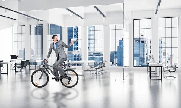 Man in business suit riding bicycle at conference hall. Businessman with megaphone looking back on bike at loft office interior with panoramic windows. Business presentation and announcement.