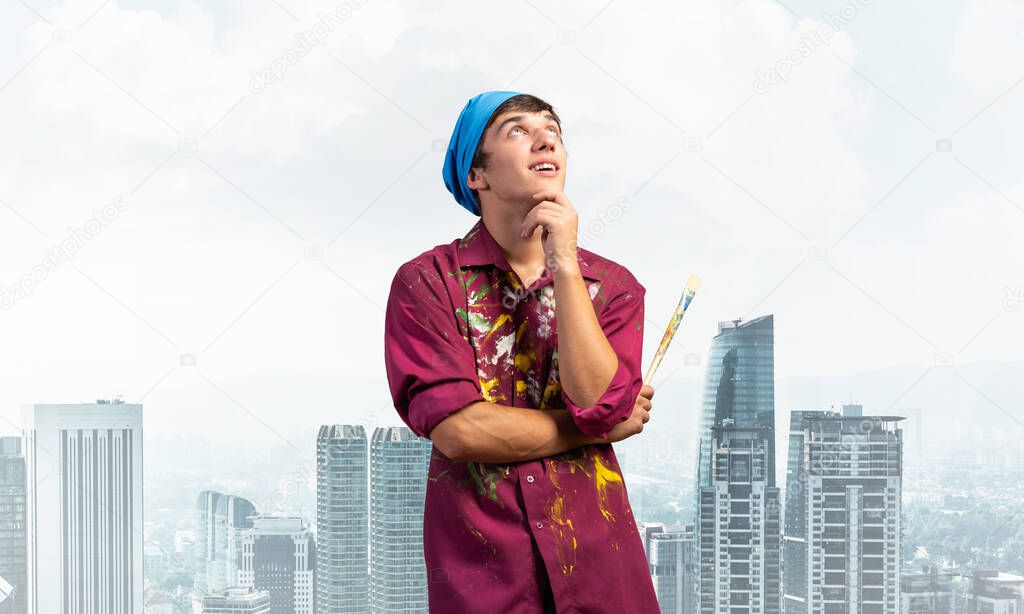 Pensive painter artist standing with folded arms on modern cityscape background. Portrait of young man looking up dreamily. Artistic occupation and creative profession. Art school student posing.