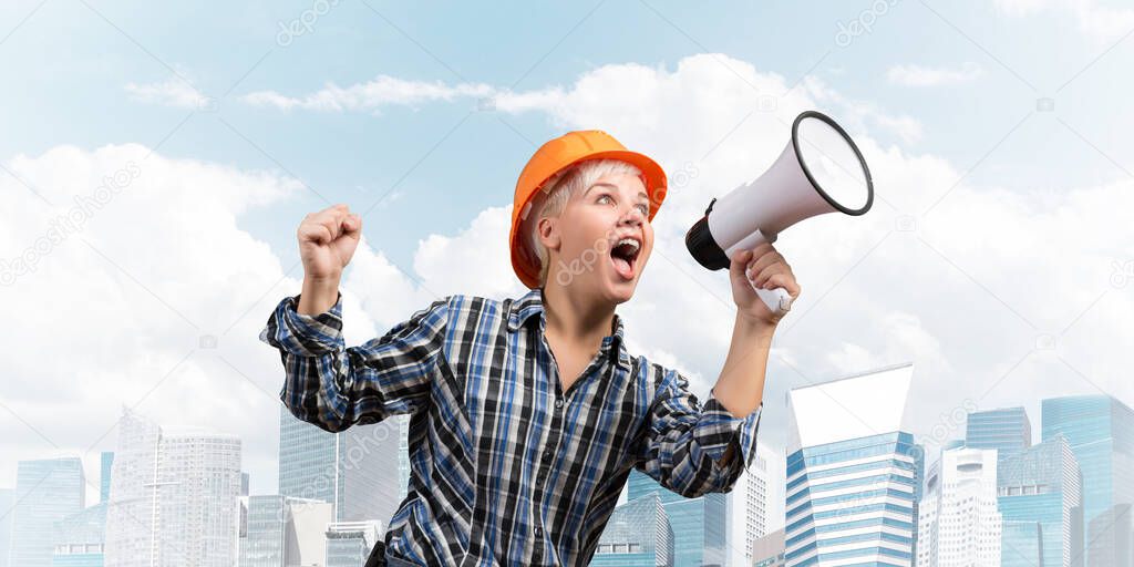 Expressive woman in safety helmet shouting into megaphone. Portrait of young emotional construction worker with loudspeaker on background of modern city. News announcement and advertisement.