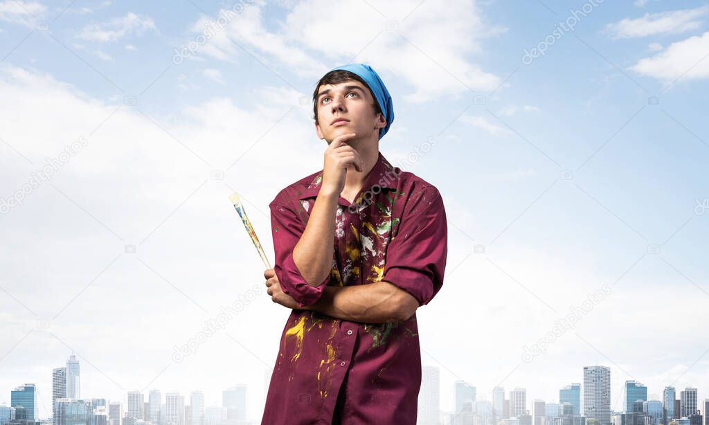 Pensive painter artist standing with folded arms on modern cityscape background. Portrait of young man looking up dreamily. Artistic occupation and creative profession. Art school student posing.