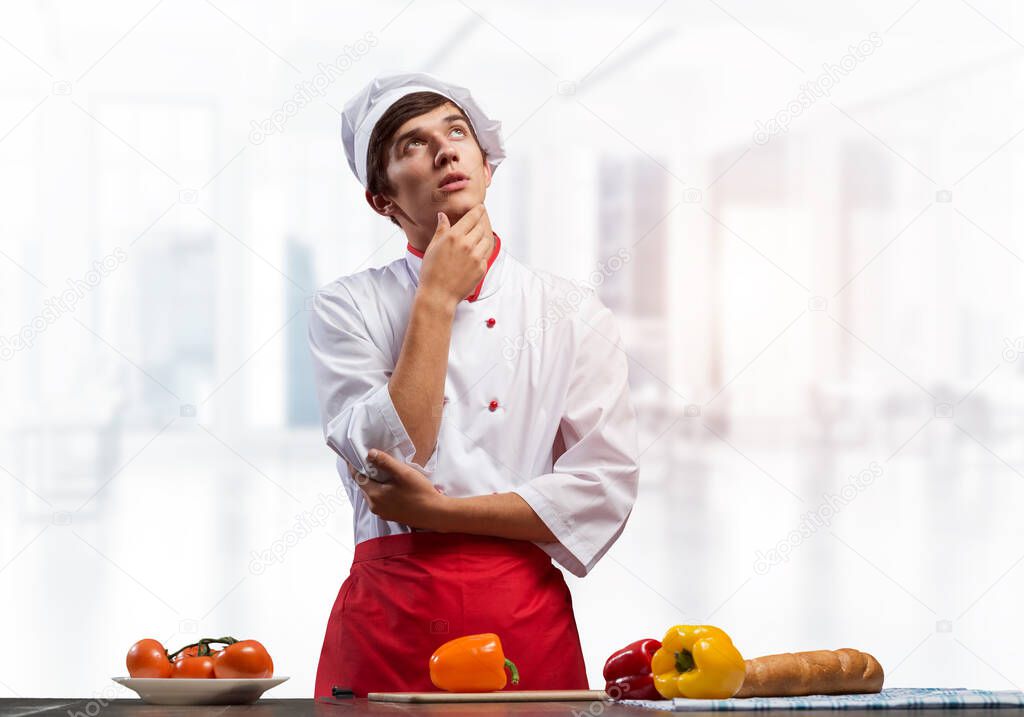 Young male chef standing with folded arms and looking up dreamily. Pensive culinary school student in white hat and red apron in light kitchen interior. Professional kitchen staff recruitment