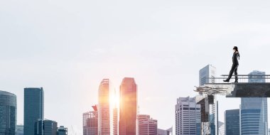 Gizli tehditlerin ve risklerin sembolü olarak beton köprüde gözü kapalı yürüyen bir iş adamı. Arka planda güneş ışığı olan şehir manzarası. 3B görüntüleme.