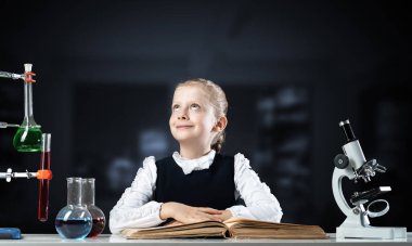 Gülümseyen küçük kız açık bir kitapla masada oturuyor. Okulda araştırma ve eğitim. Kütüphanede okuyan mutlu liseli kız. Okuldaki zeki kız karanlık arka planla ilgili kitap okuyor..