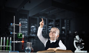 İlköğretim fen sınıfında masa başında oturan küçük bir bilim kadını. Okulda araştırma ve eğitim. Mikroskop, matara ve test tüplü kimya laboratuvarı. Mutlu liseli kız işaret ediyor