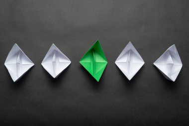 Siyah arka plan üzerinde kağıt gemi sırası. Bireysel motivasyon ve yön. Yaratıcı yenilik ve liderlik. Düz beyaz tekne grubunda yeşil origami tekne yatıyordu. Sosyal pazarlama düzeni.