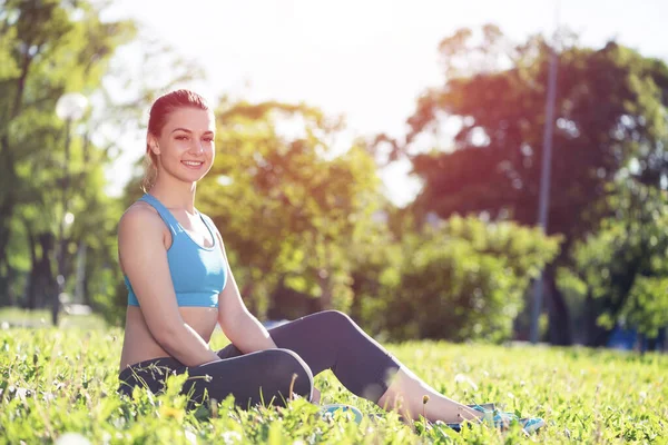公園でリラックスしてスポーツウェアの美しい笑顔の女の子 トレーニングの後 緑の芝生の上に座って若い女性 晴れた夏の日に瞑想屋外 朝の演習と健康的なライフスタイル — ストック写真