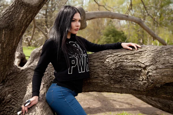 Hezká mladá brunetka žena nosí džíny a černou košili s velkým zrcadlem brýle pózuje u stromu v parku — Stock fotografie