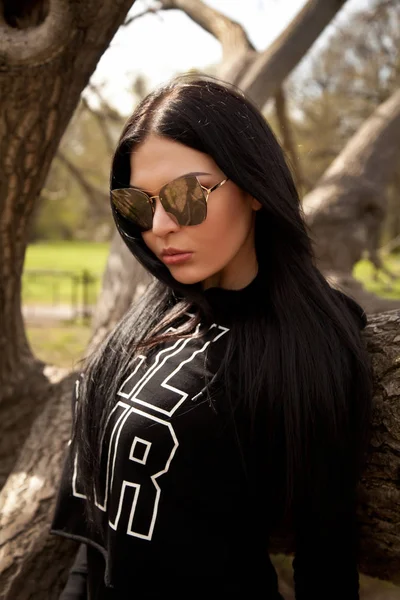 Mooie jonge brunette vrouw het dragen van jeans en zwarte shirt met een grote spiegel zonnebril poseren in de buurt van de boom in het park — Stockfoto
