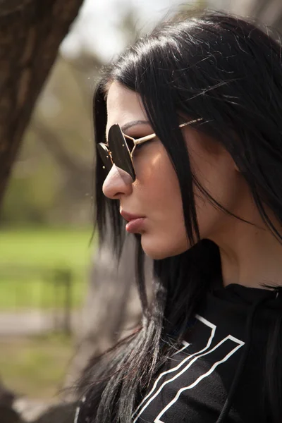 Mooie jonge brunette vrouw het dragen van jeans en zwarte shirt met een grote spiegel zonnebril poseren in de buurt van de boom in het park — Stockfoto