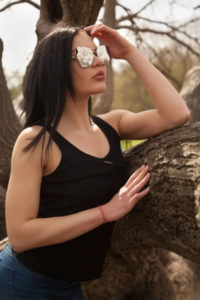 공원에서 나무 근처 포즈 큰 미러 선글라스와 청바지와 검은 셔츠를 입고 아주 젊은 갈색 머리 여자 — 스톡 사진