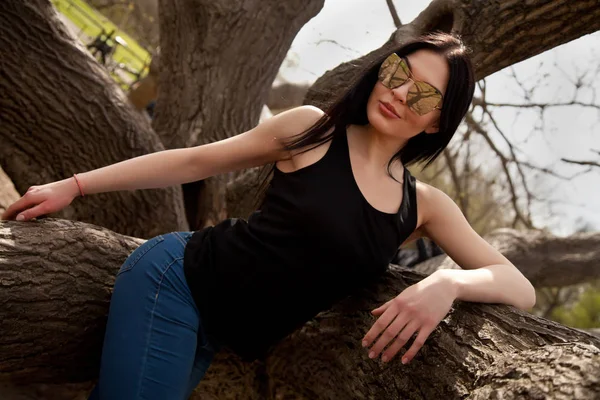 Pretty young brunette woman wearing jeans and black shirt with big mirror sunglasses posing near the tree in the park — Stock Photo, Image