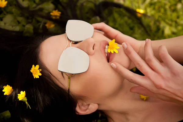 Krásná mladá brunetka s stříbrné zrcadlo brýle ležící na trávě se žlutými květy v létě — Stock fotografie