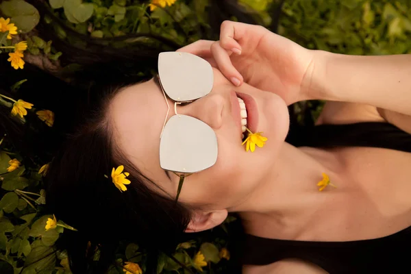 Mooie jonge brunette met zilveren spiegel zonnebril liggend op het gras met gele bloemen in de zomer — Stockfoto