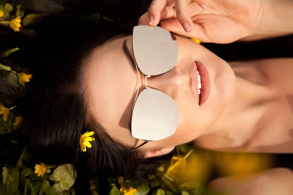 Красивая молодая брюнетка в серебряных зеркальных солнцезащитных очках, лежащих на траве с желтыми цветами летом — стоковое фото